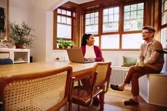 Female sitting at a table with laptop talking to male with coffee mug at home
