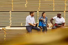 Three colleagues having a laugh during a lunch break while sitting on a pile of wood.
